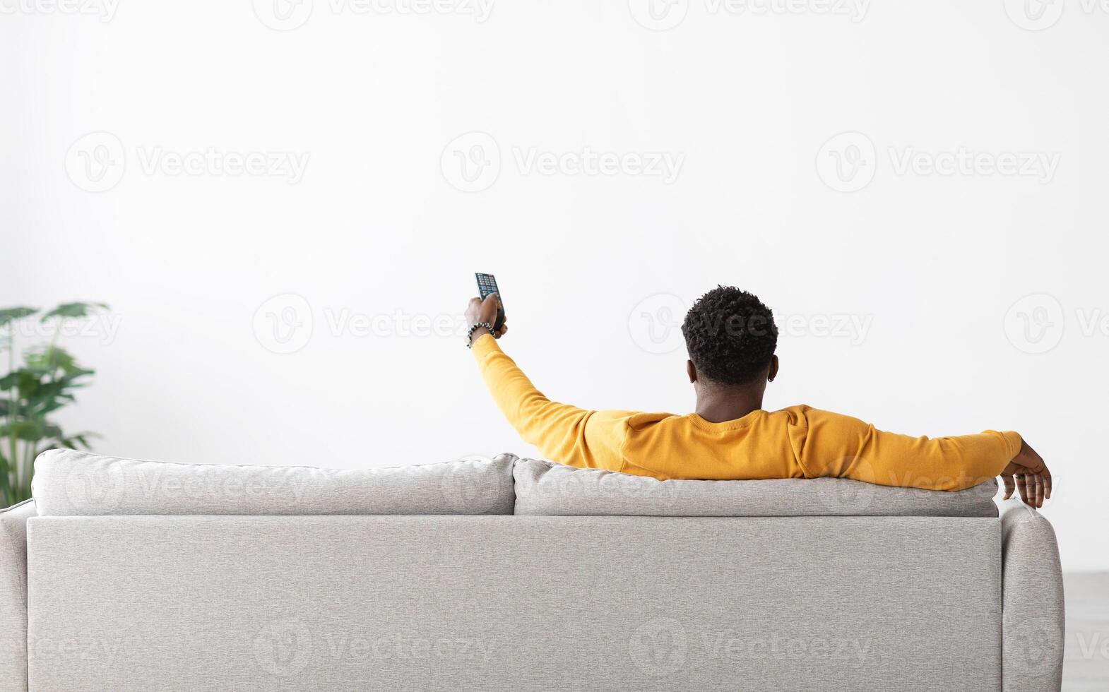 Back view of black man in yellow sitting on couch with remote from TV, pointing towards empty white wall, african american guy turning on air conditioner, home interior, copy space photo