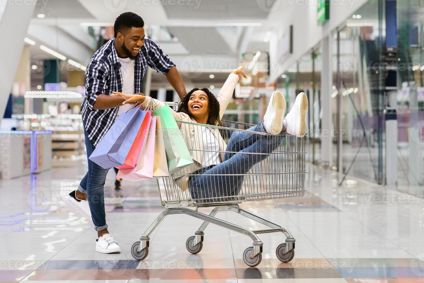 Cheerful black man pulling shopping trolley with his girlfriend inside, having fun while making purchases together in modern department store, fooling and laughing, enjoying seasonal sales, copy space photo