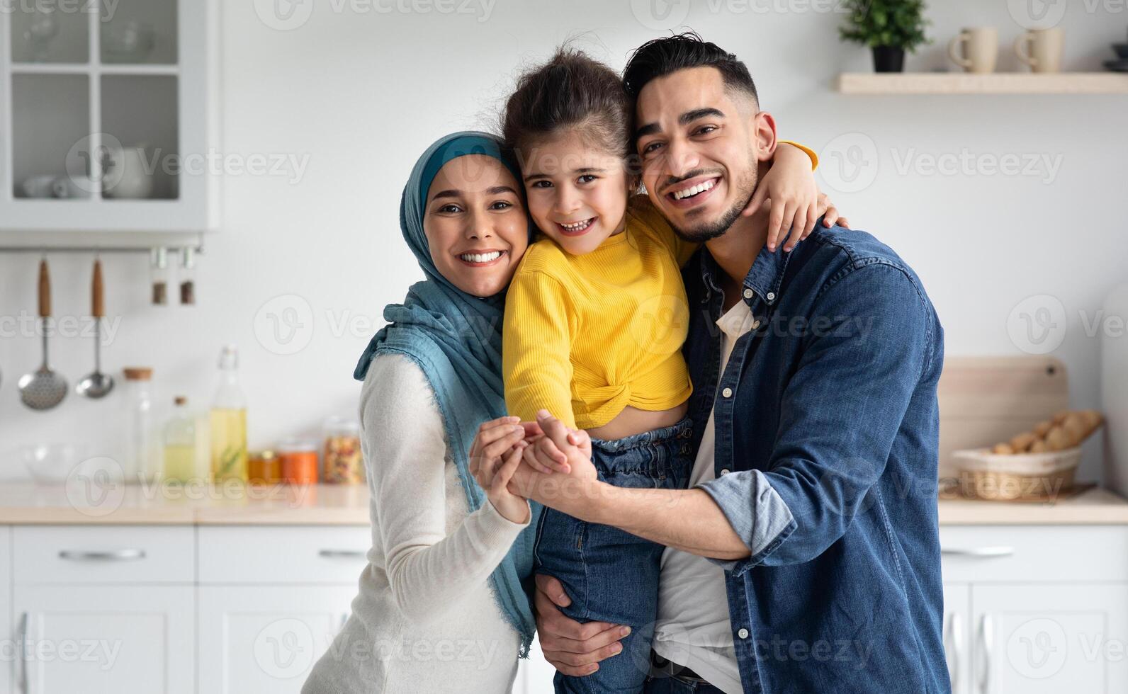 Portrait Of Happy Muslim Family Of Three With little Daughter Posing In Kitchen, Cheerful Islamic Father, Child And Mother In Hijab Embracing And Smiling At Camera, Bonding At Home Together photo