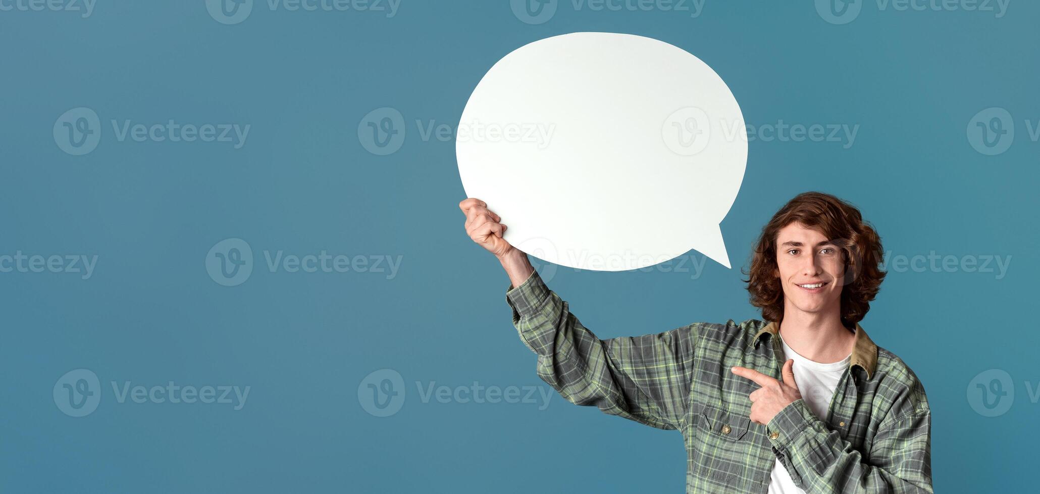 Communication concept. Cheerful curly guy pointing at speech bubble on color background photo