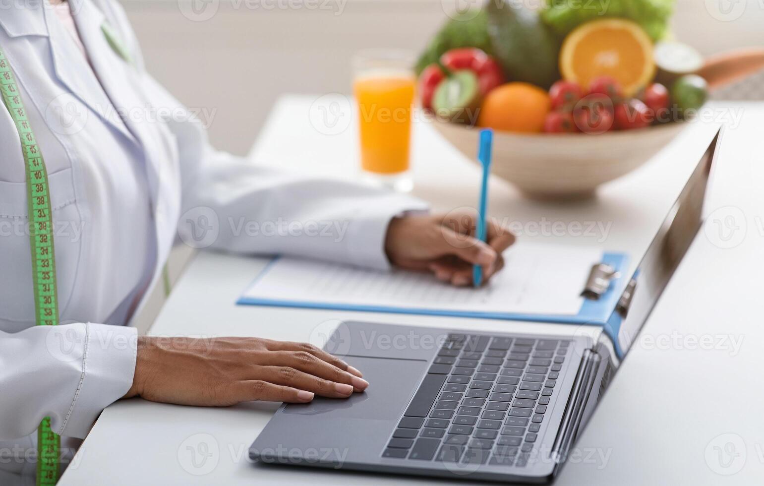 Cropped of black woman nutritionist working online, using laptop and writing down client history, clinic interior photo