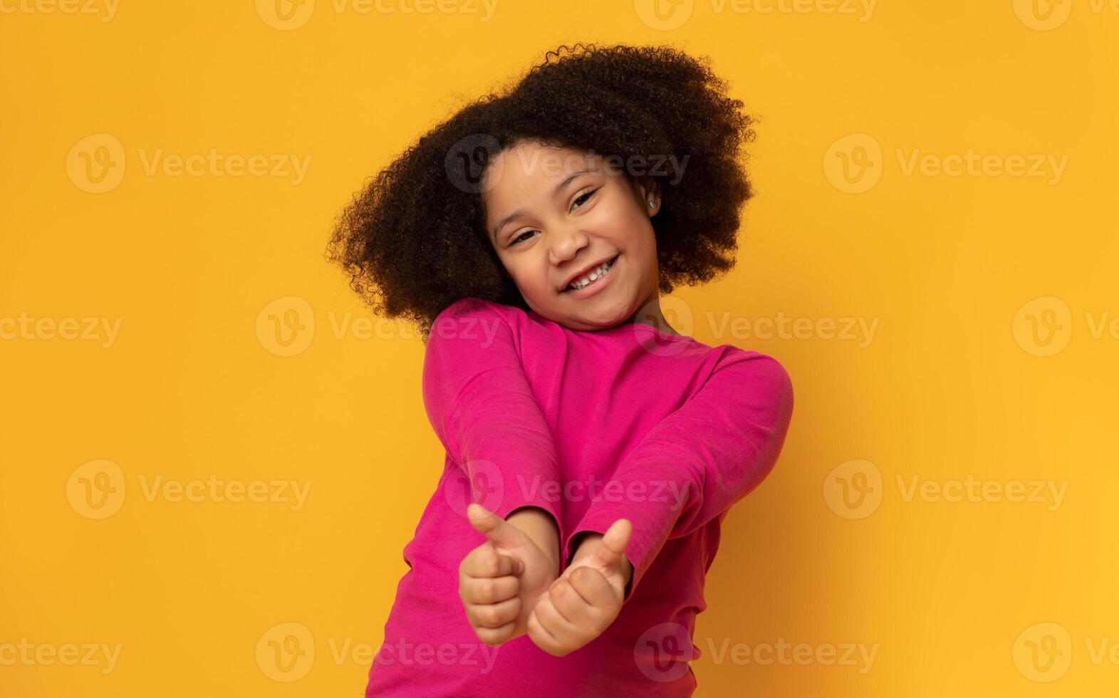 Everything is great. Positive cute little girl gesturing thumbs up and smiling at camera, yellow studio background with empty space photo