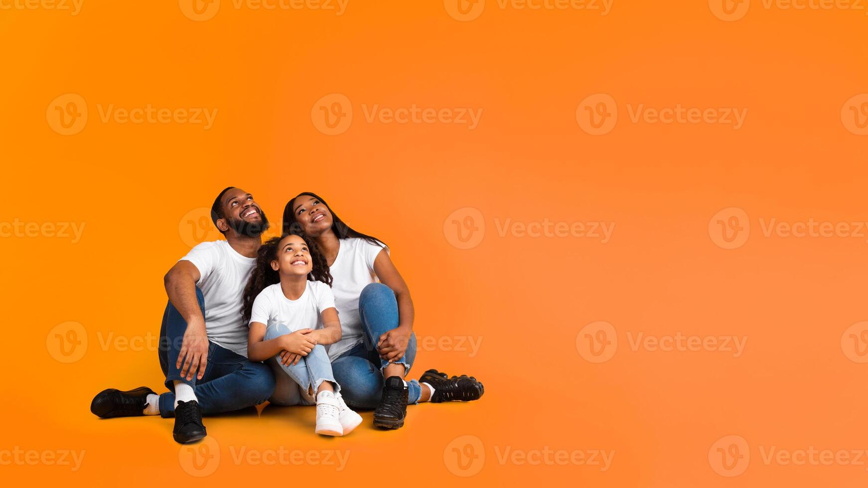 Promo And Advertisement. Smiling African American Family Of Three People Sitting On The Floor, Looking Up At Free Empty Copy Space Isolated On Yellow Orange Studio Background, Banner, Panorama photo