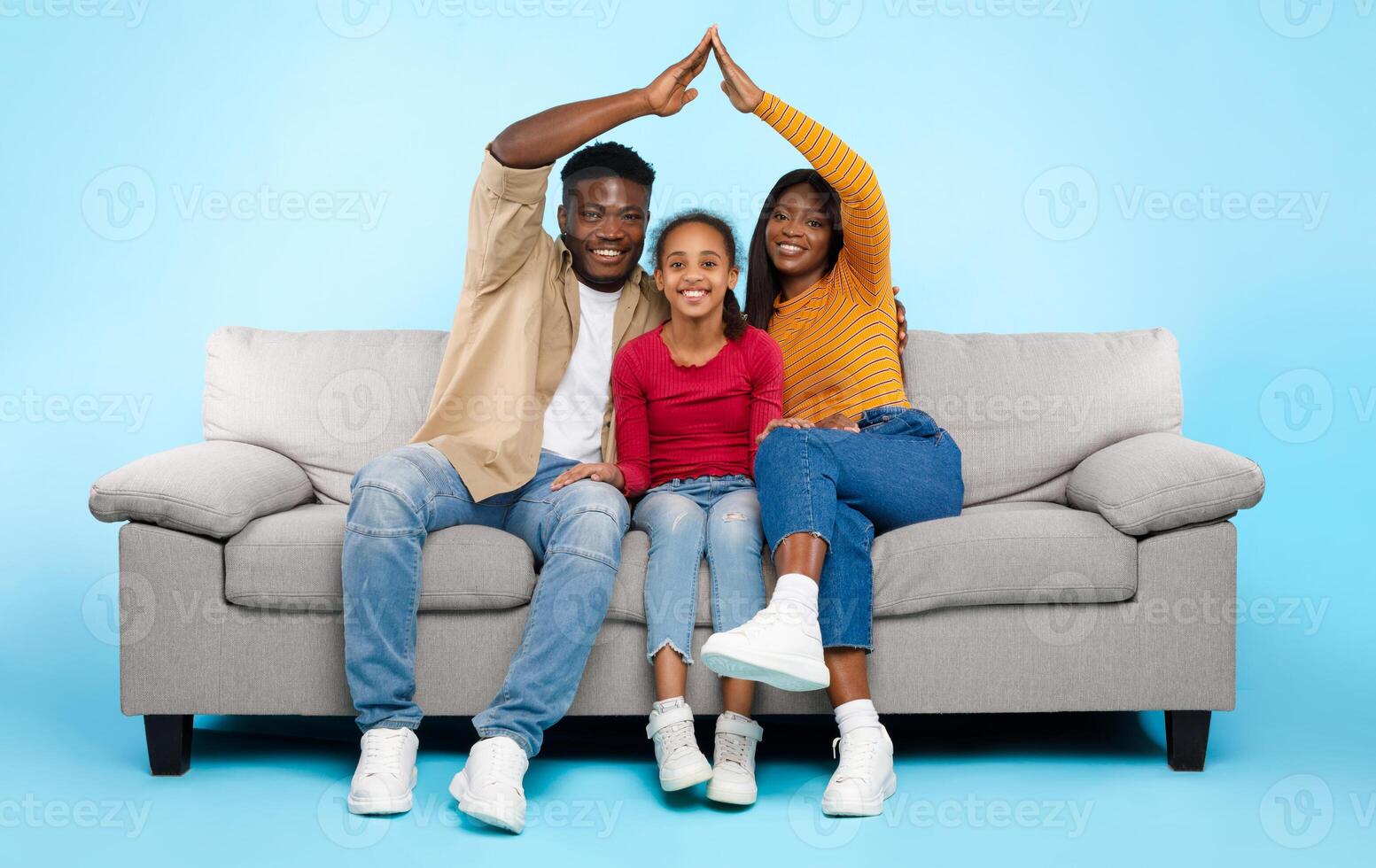 Family Care, Protection And Insurance Concept. Portrait of smiling young African American parents making symbolic roof of hands above their happy daughter sitting on sofa couch at blue studio wall photo