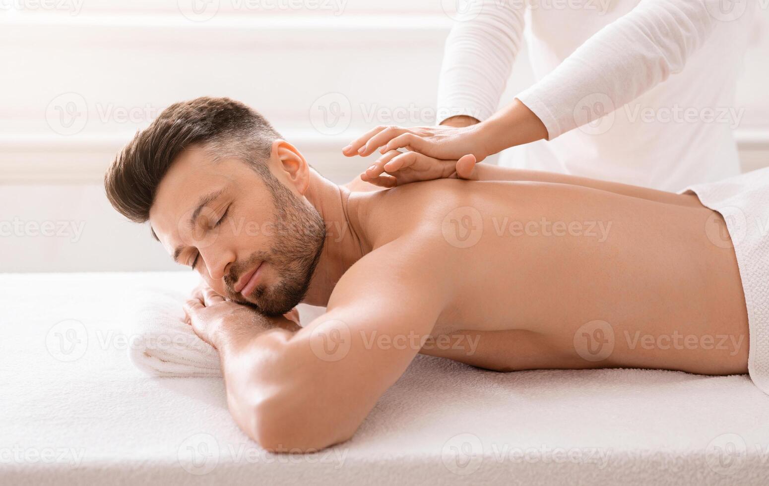 Side view of relaxed handsome man having body massage at spa salon. Middle aged bearded man attending modern male spa, laying on massage table, getting healing body procedure, enjoying his day at spa photo