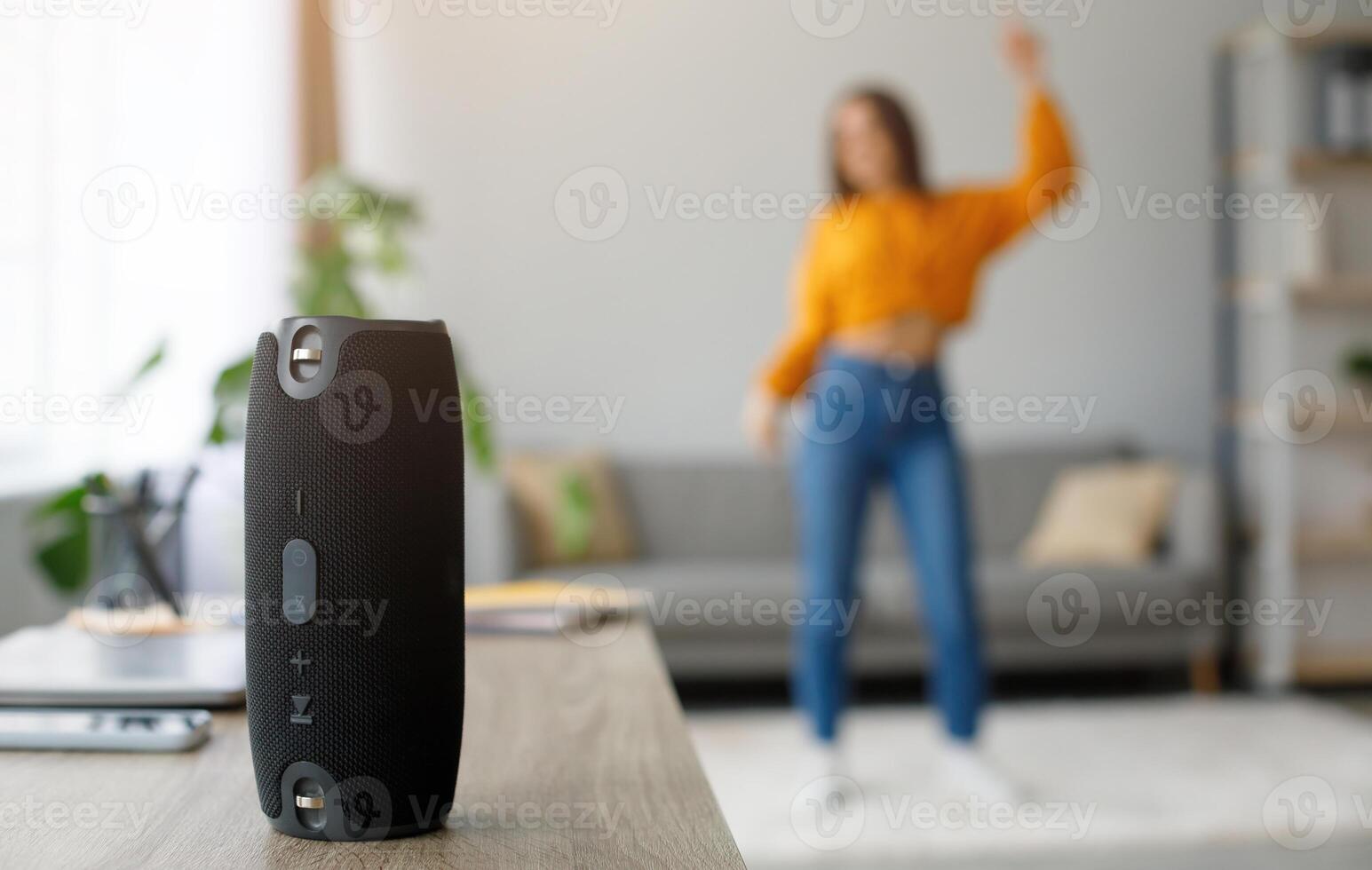 Millennial woman dancing to favorite music at home, selective focus on portable wireless speaker, free space. Young female moving to popular song, using modern stereo system indoors photo