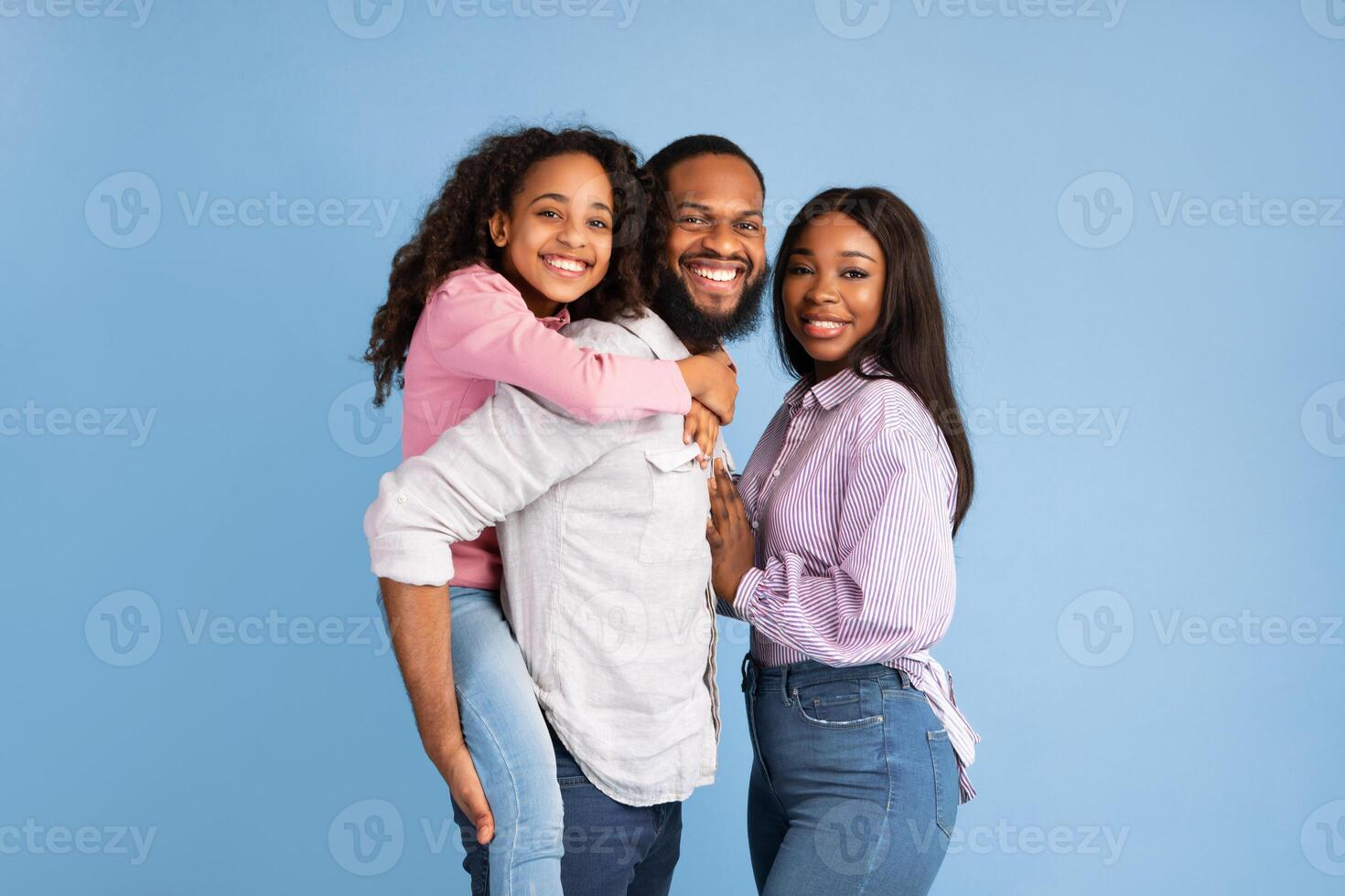 Time With Family. Portrait of cheerful African American dad, mum and daughter posing looking at camera standing isolated over blue studio wall. Happy father carrying adolescent girl on back photo