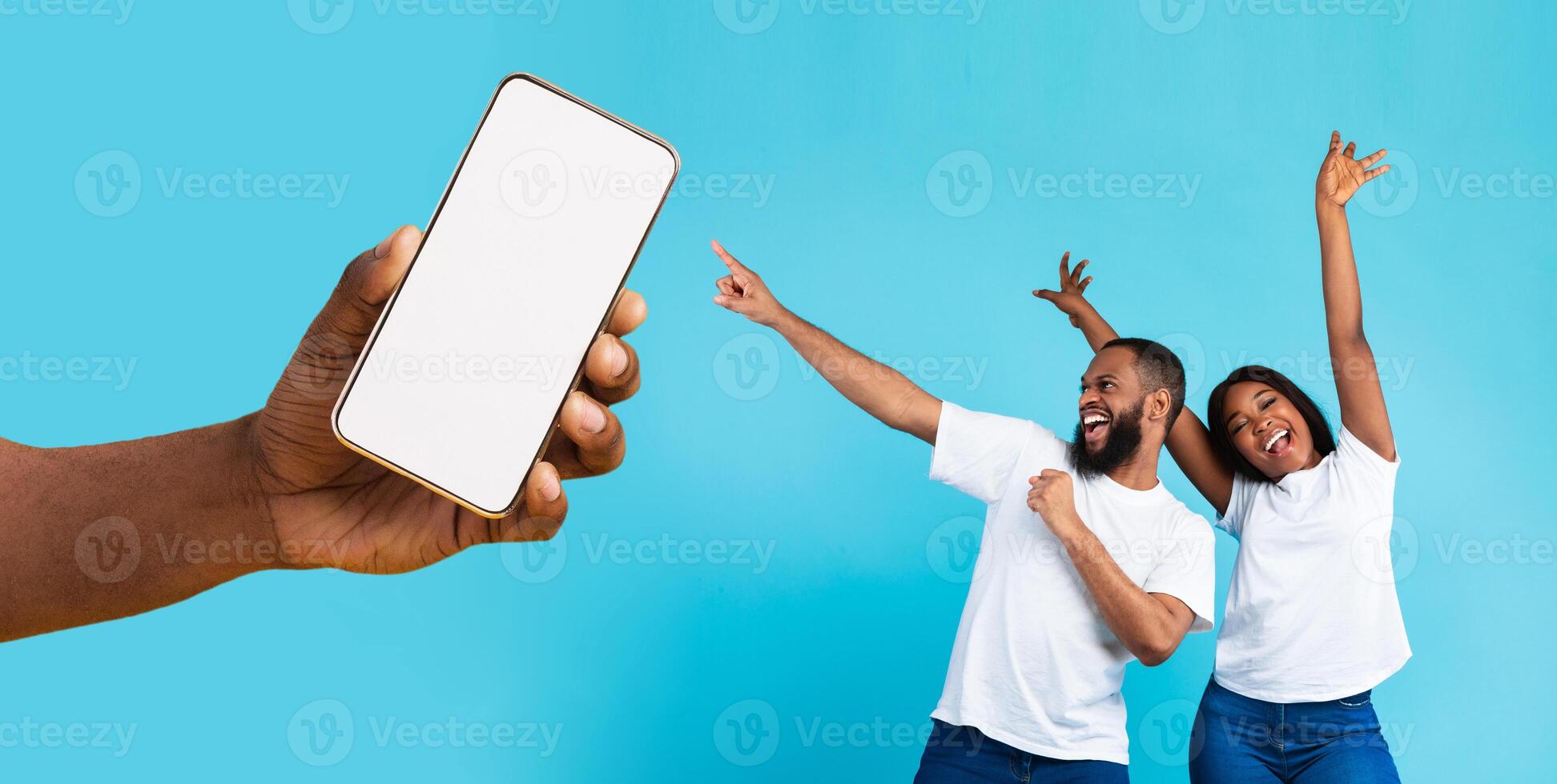Great Offer. Cheerful African American couple dancing and pointing at giant cell phone showing blank space for mock up, celebrating win, hand holding white empty screen, blue studio background wall photo