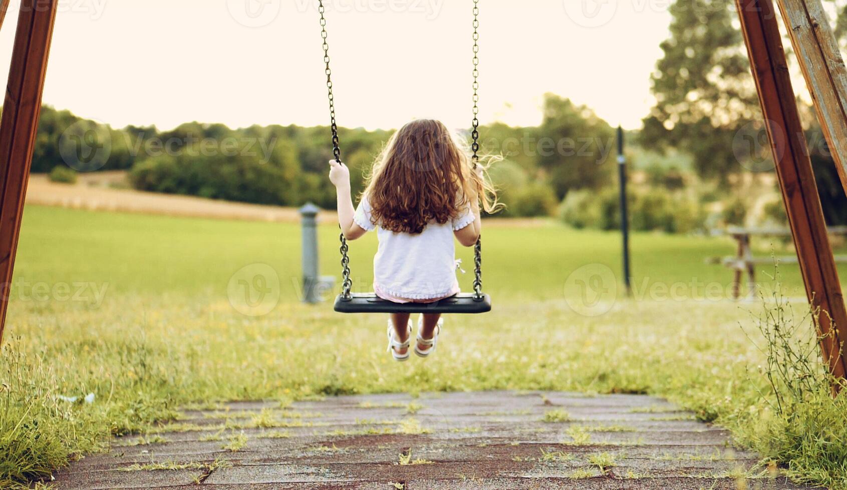 Back view of a little long-haired girl, swinging on a swing in a green meadow. Happy carefree childhood of a cute child. photo