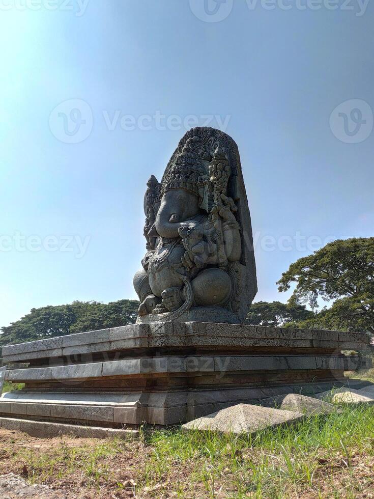 A majestic stone sculpture of Lord Ganesha, intricately carved with a large head and crown, holds an axe and modak. Seated against a clear sky and green trees, it reflects rich Hoysala heritage. photo