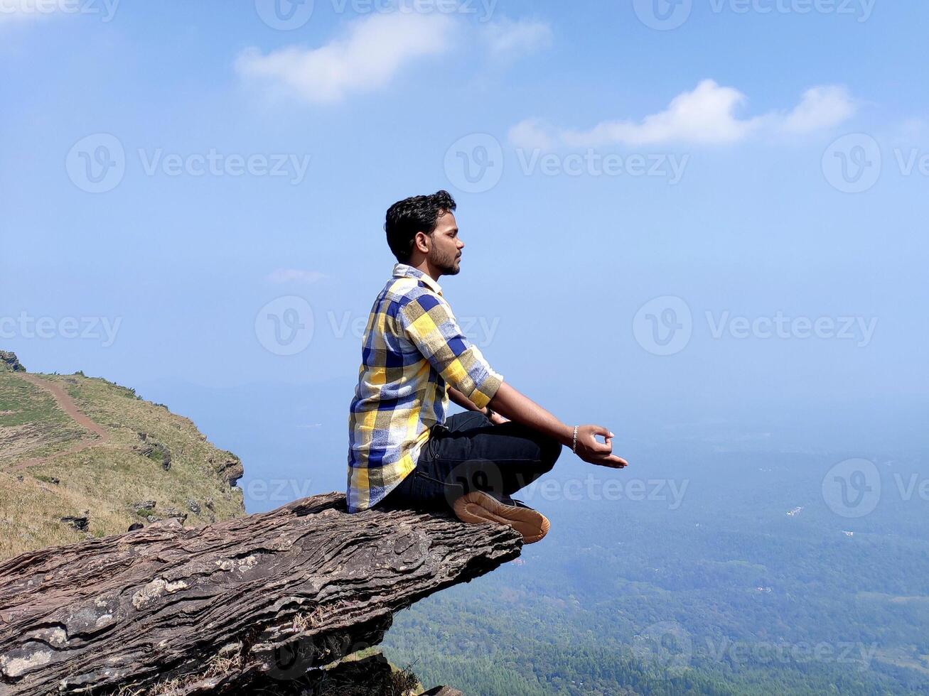 A man meditates on a cliff edge, cross-legged with hands in mudra, overlooking a vast green valley and blue sky. Wearing a checkered shirt, he embodies calm, focus, and harmony in natures beauty. photo