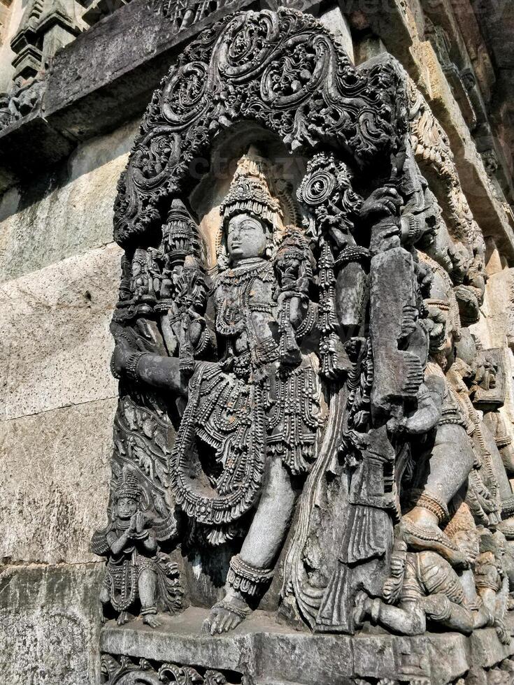 A sculpture of Vamana, fifth avatar of Vishnu, adorns a wall panel at Belur's Chennakeshava Temple. Vamana stands in a dynamic pose with one leg raised, detailed in intricate garments and jewelry. photo