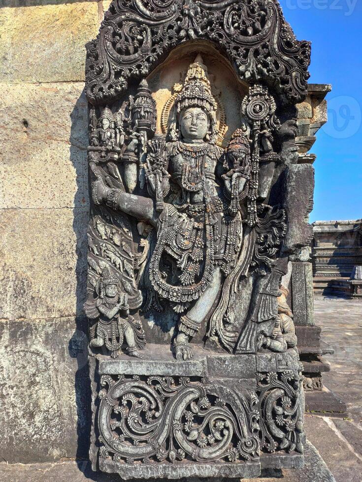 A detailed stone carving of Vishnu, adorns a wall at Chennakeshava Temple. Vishnu stands dynamically, right leg raised, displaying intricate details in garments, jewelry, and physique. photo