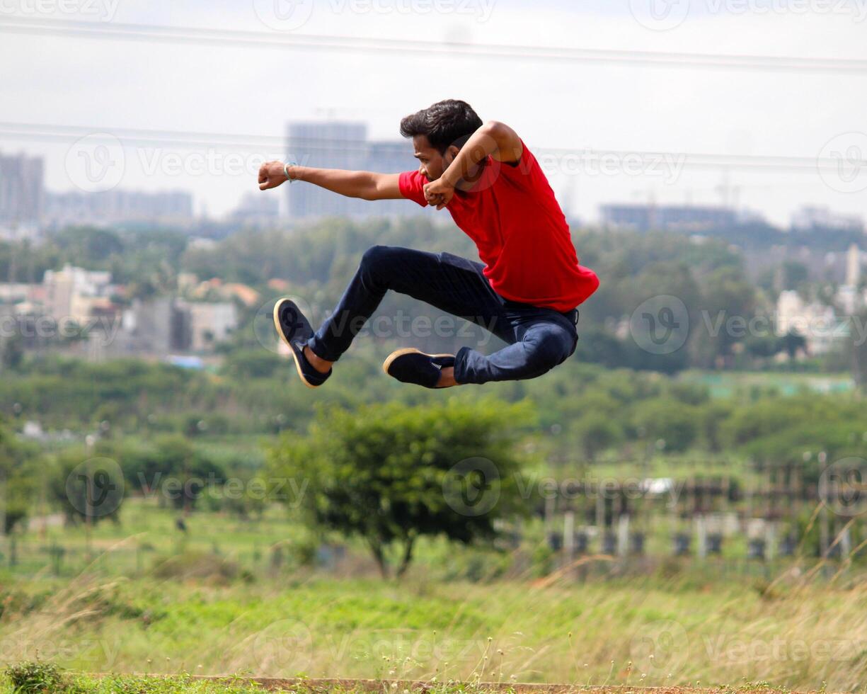 An energetic young man in mid-air performs a martial arts-style jump. Dressed in red and dark pants, he strikes a powerful pose against a grassy field and blurred cityscape, symbolizing energy. photo