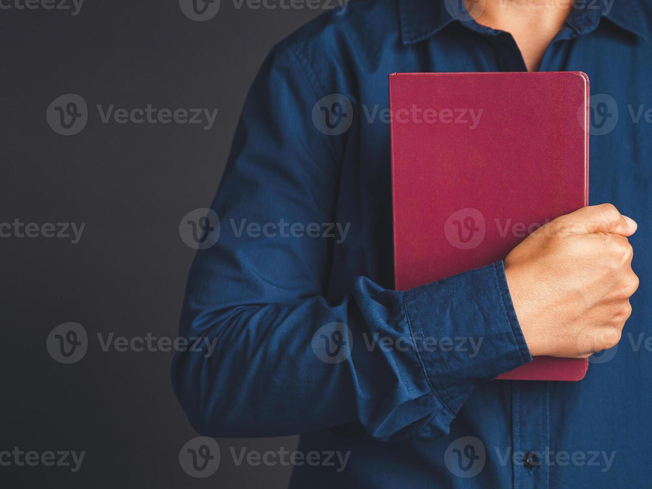 Close view of man in blue shirt holding a book with an empty red cover in hand while standing on a gray background photo