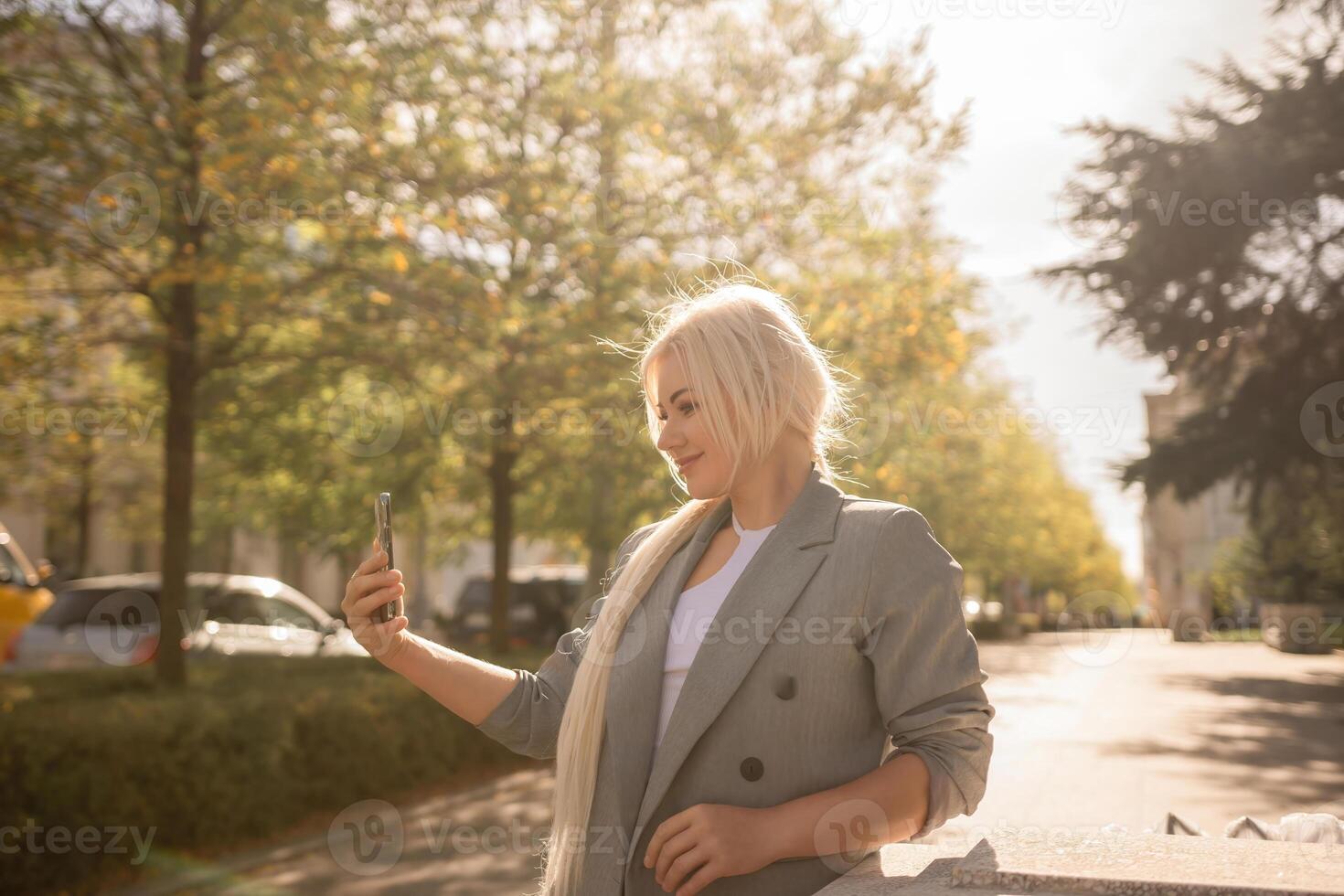 A woman is taking a picture of herself with her cell phone. She is wearing a gray jacket and scarf. The scene is set in a city with trees and cars in the background. photo