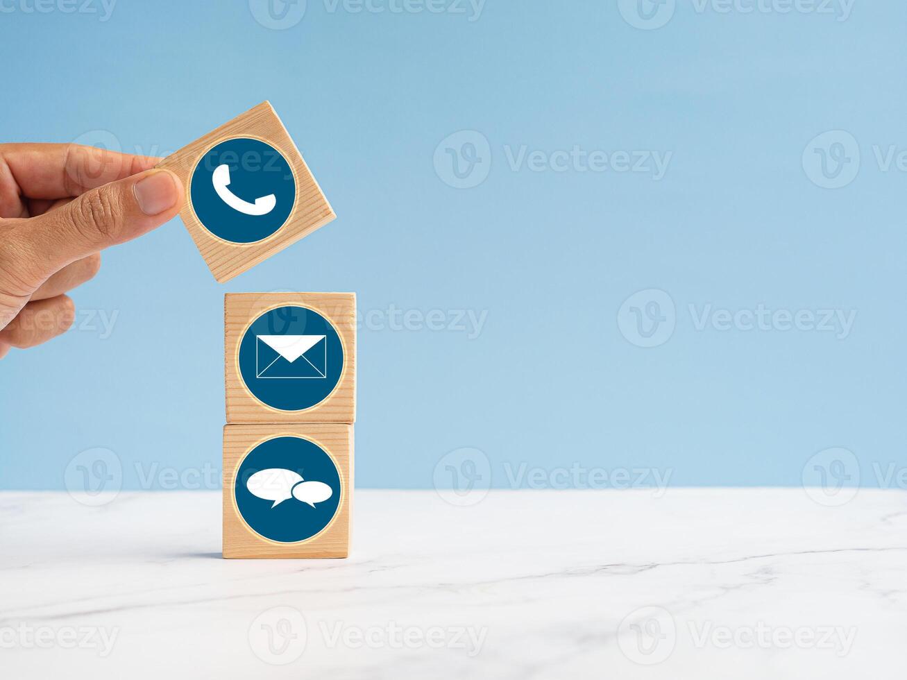 Wooden cubes with symbols of telephone, email, and speech bubble over a marble floor against a light blue background. Contact us concept photo