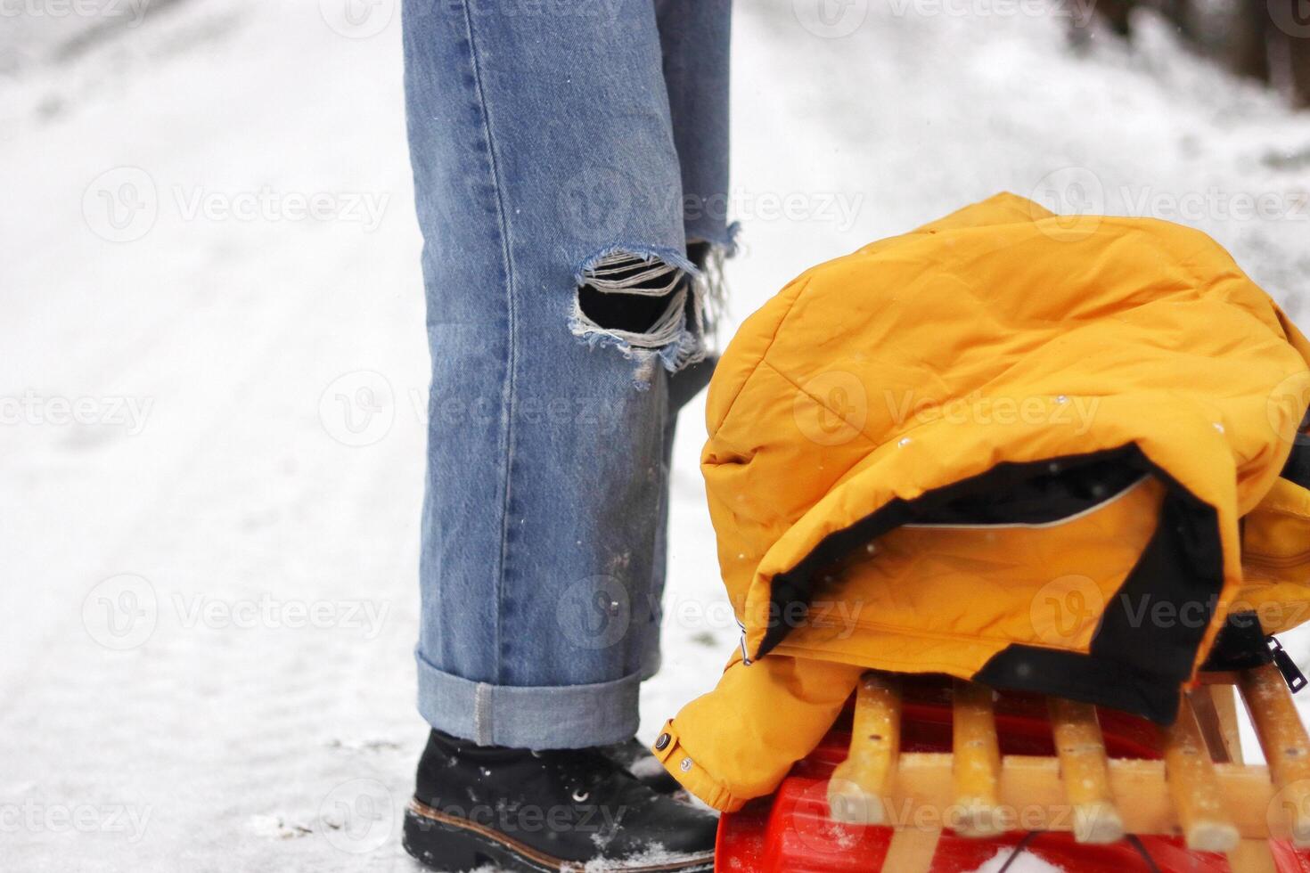 Winter scene with person in ripped jeans and black boots standing near a wooden sled with a yellow jacket on top. Snowy forest path on a cold winter day. Copy space photo
