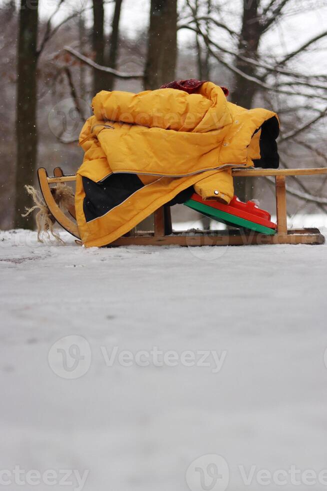 Clolseup of a wooden sled with a warm yellow jacket on a snowy ground in the woods, depicting a winter scene and peaceful retreat into a snow-covered forest. Copy space photo