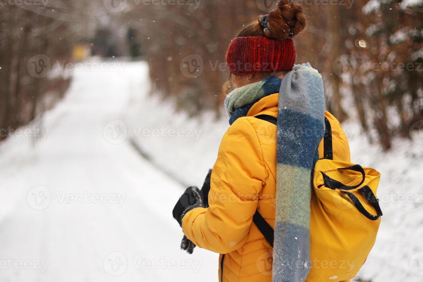 Winter scene with a young woman dressed warmly in a yellow jacket, scarf and red knit headband, walking down a snow covered path through a quiet forest landscape. photo
