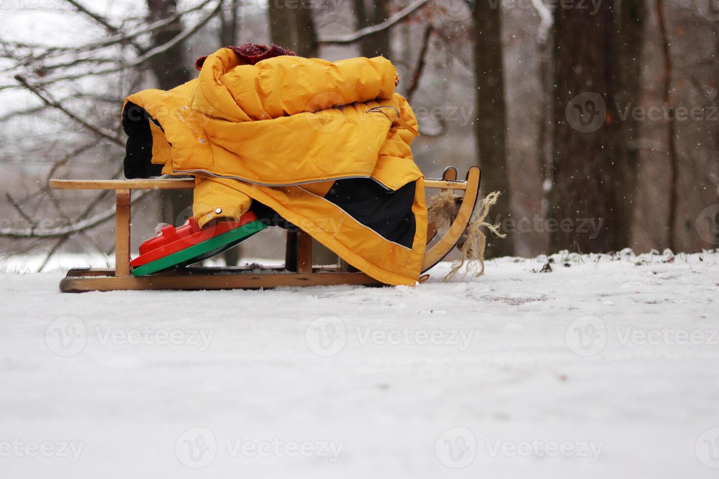 Clolseup of a wooden sled with a warm yellow jacket on a snowy ground in the woods, depicting a winter scene and peaceful retreat into a snow-covered forest. Copy space photo