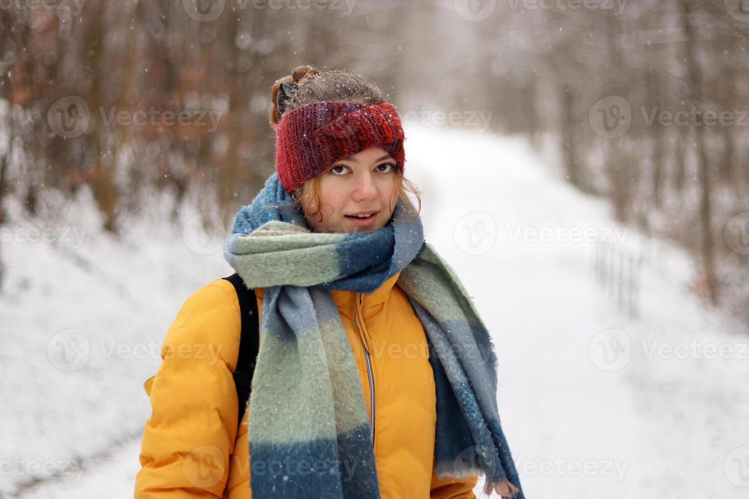 Closeup portrait of a smiling young woman wearing a red knit headband, scarf, and yellow jacket on a cold winter day. Girl dressed warmly surrounded by a blurred snowy forest. photo