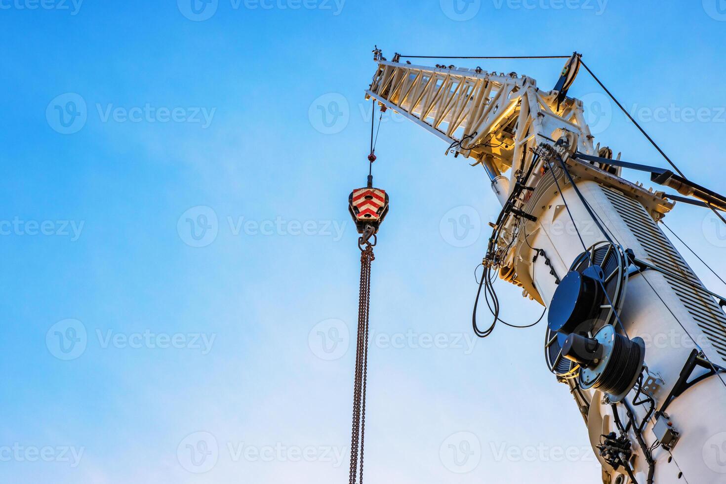 A large construction crane with cables extending against a clear blue sky, White crane arm and hanging cables captured from below, illustrating industrial work and heavy lifting. Copy space photo