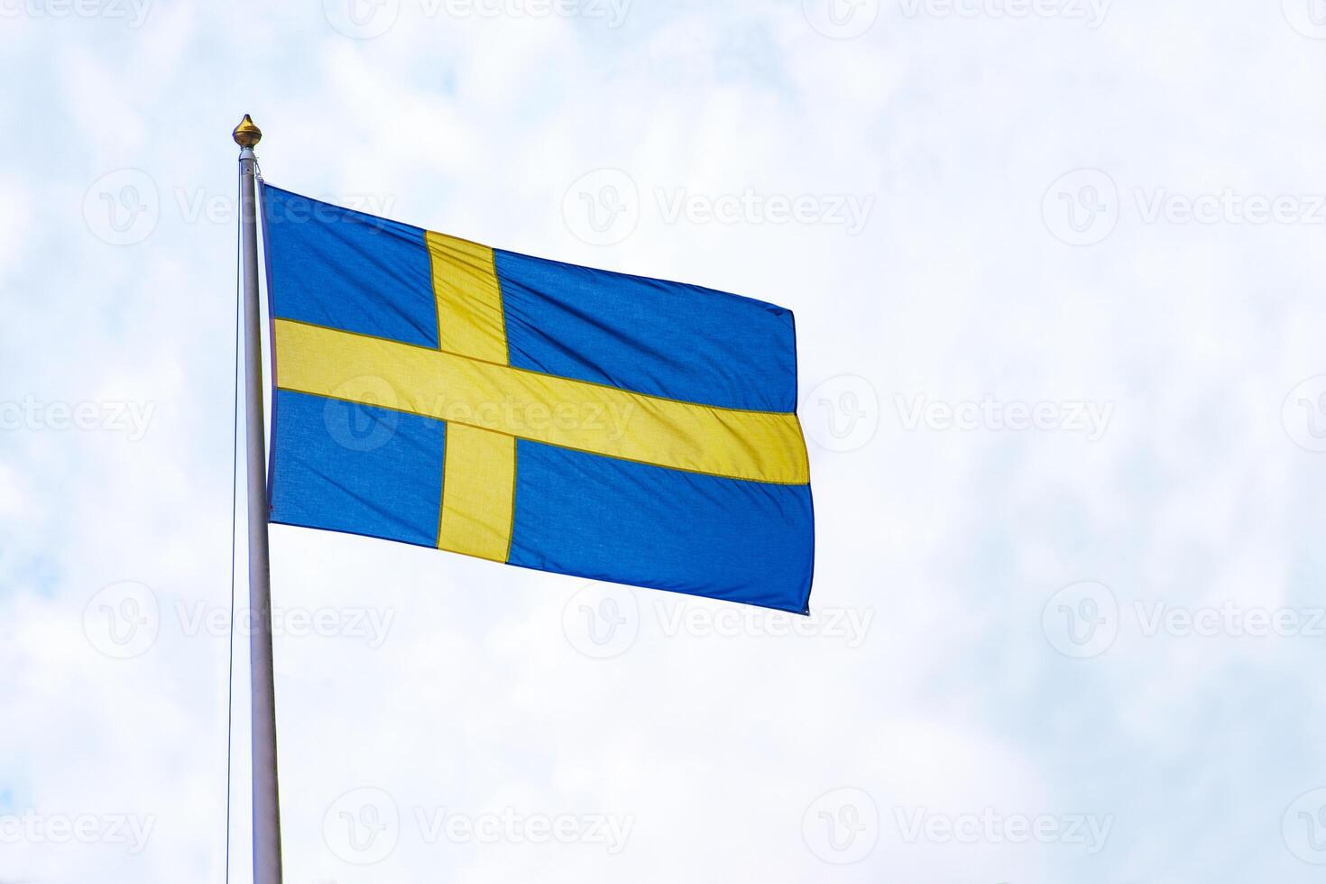 Swedish flag waving against a bright sky. The national flag of Sweden, featuring a golden cross on a blue background, flying on a flagpole under a partly cloudy sky. Copy space photo