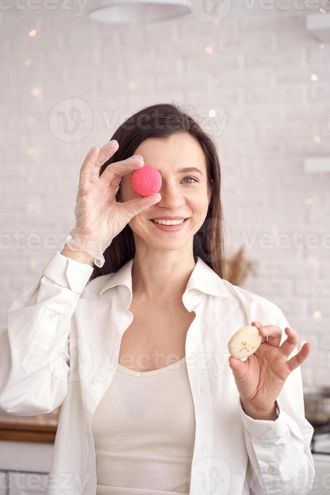 Smiling woman holding macarons in a modern kitchen setting with fairy lights in the background. photo