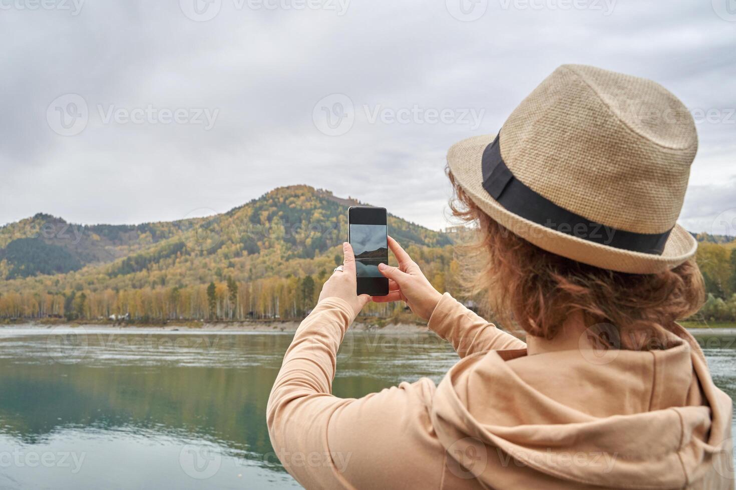 Traveler captures scenic mountain view with smartphone while wearing hat by peaceful lake. photo
