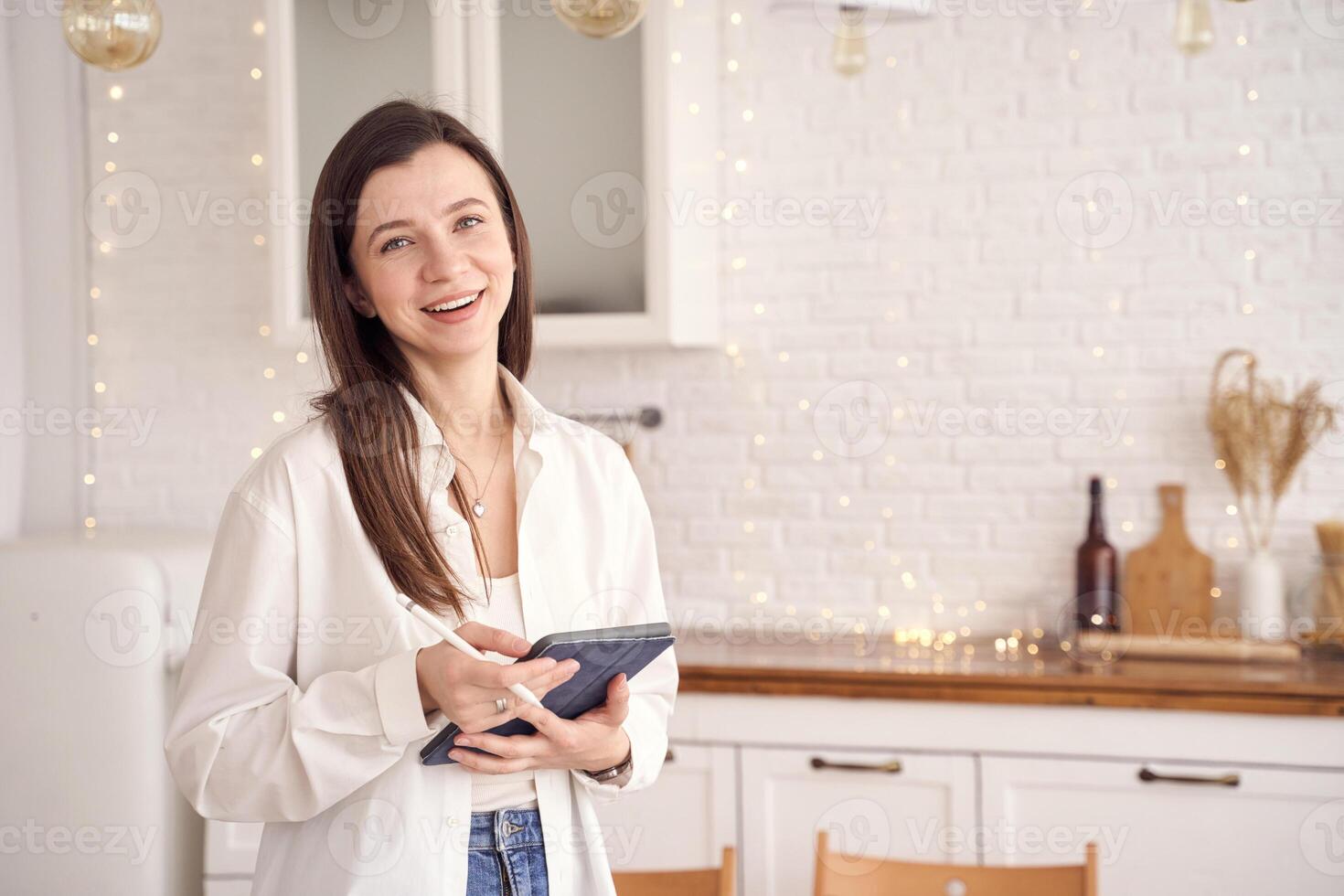 Smiling woman using tablet in cozy kitchen with warm lighting and string lights. photo