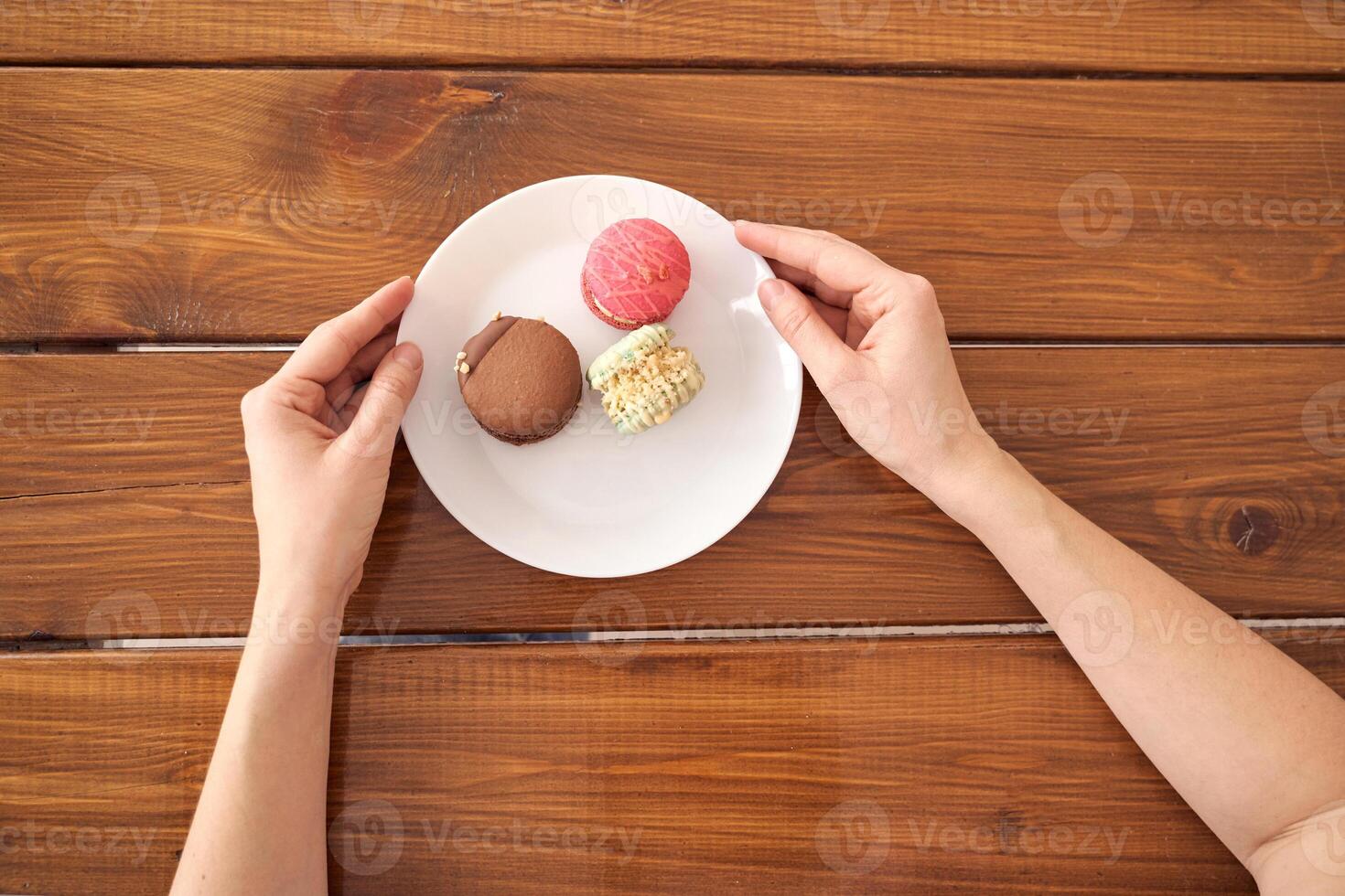 Hands holding white plate with assorted macarons on rustic wooden table background. photo