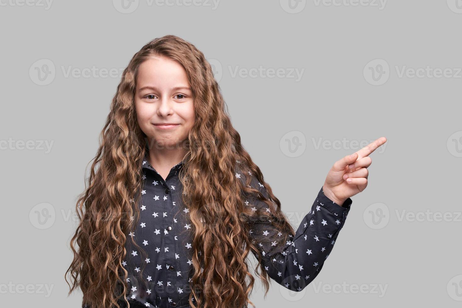 Confident young girl smiling and pointing to the side with long curly hair in starry patterned shirt photo