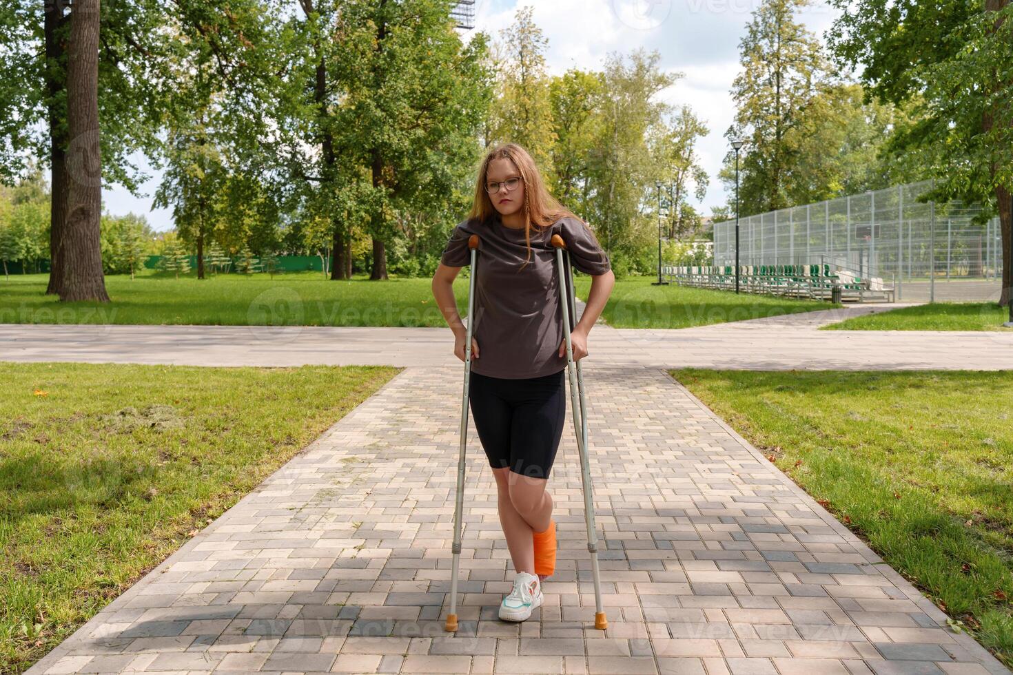 Crutches supporting her,teenage girl with broken leg in cast moves through summer park,bright, nurturing environment emphasizing her resilience and ongoing process of recovery photo