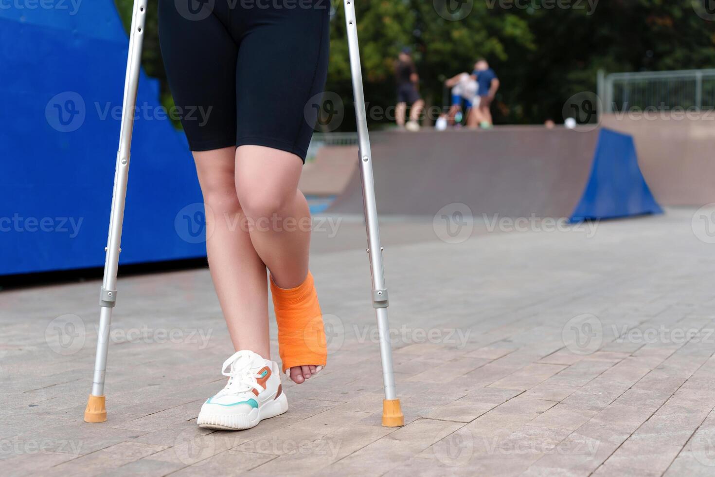 plastered female leg in an orange bandage in the background of a summer skate park, crutches for balance and mobility aid photo