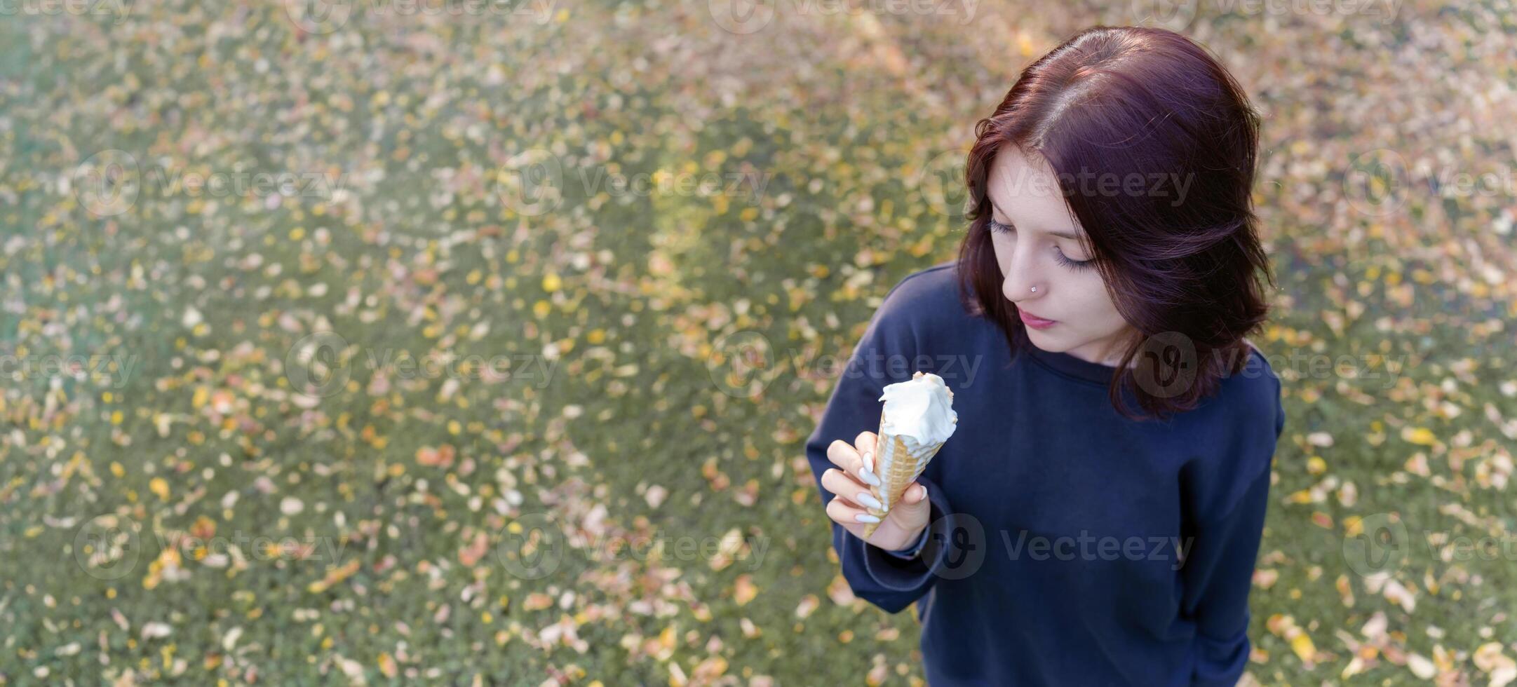 Above is a view of a young girl with an unusual appearance seductively licking cream ice cream in a waffle cup, surrounded by yellowing foliage and falling leaves photo