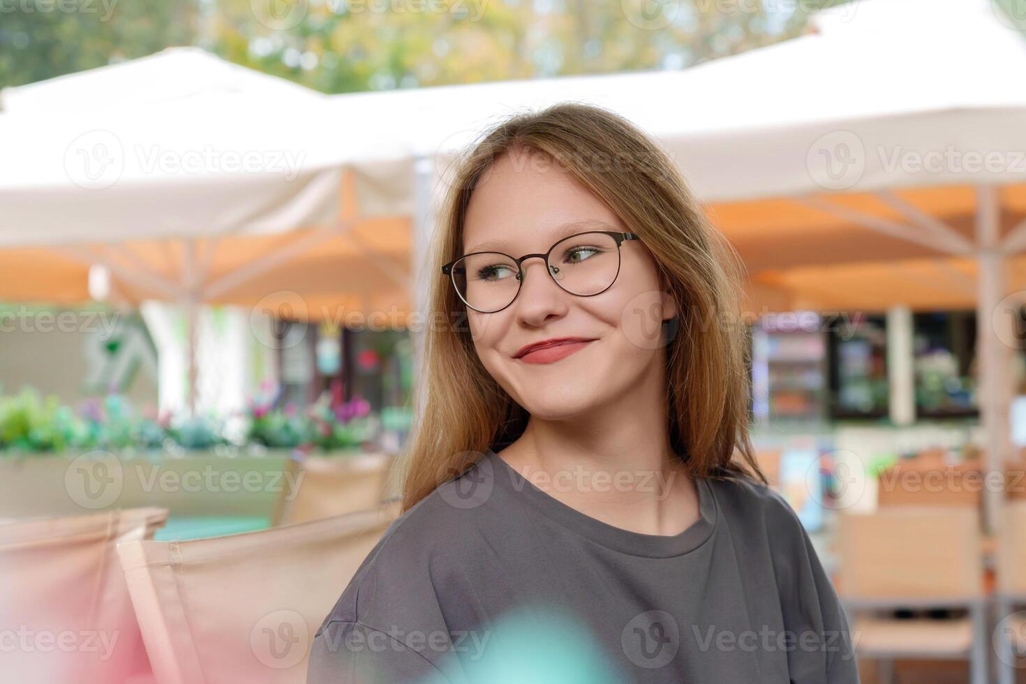 radiant, fair-haired teenage girl breaks into a warm smile as she sits at a table in a lively summer cafe, her joyful expression exuding a sense of relaxed contentment photo