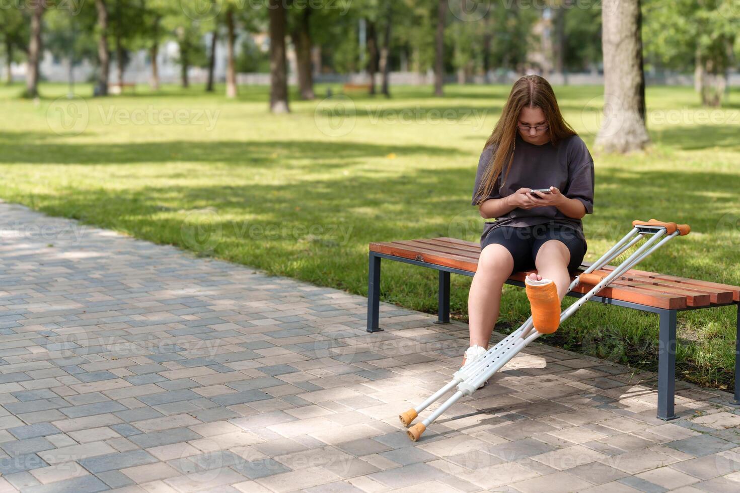 young girl sits on a bench in a sunny park and uses her smartphone while placing her broken leg on the crutches lying nearby photo