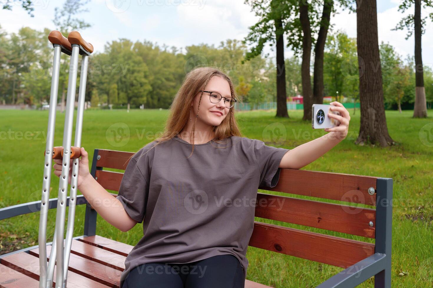 Young female with a casted leg pausing on a bench to take a self-portrait, preserving a moment of her rehabilitation journey and the challenges faced in the tranquil summer setting photo