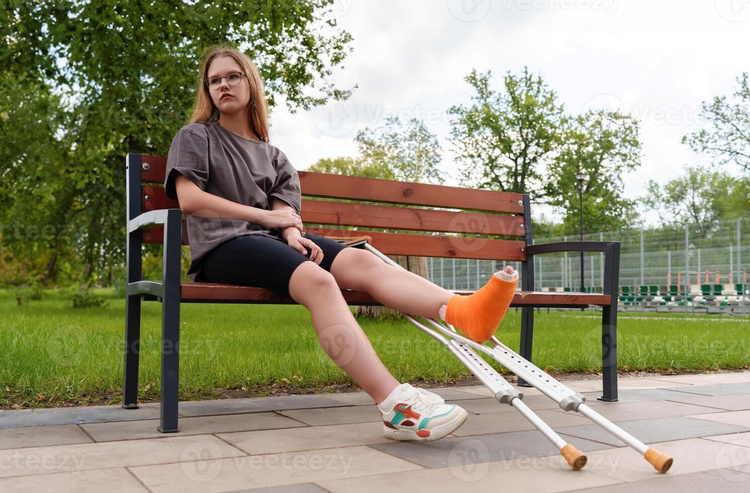 Casual-dressed female adolescent with cast on leg appears disheartened while seated on bench in summer park, conveying emotional and physical challenges during recovery. medical, disability, lifestyle photo