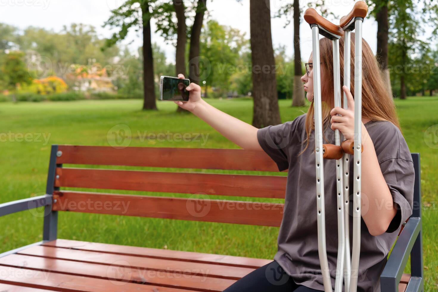 Teenage girl with a leg cast seated on a park bench, capturing a bittersweet selfie as a memento of her injury and the time lost during the recovery process photo