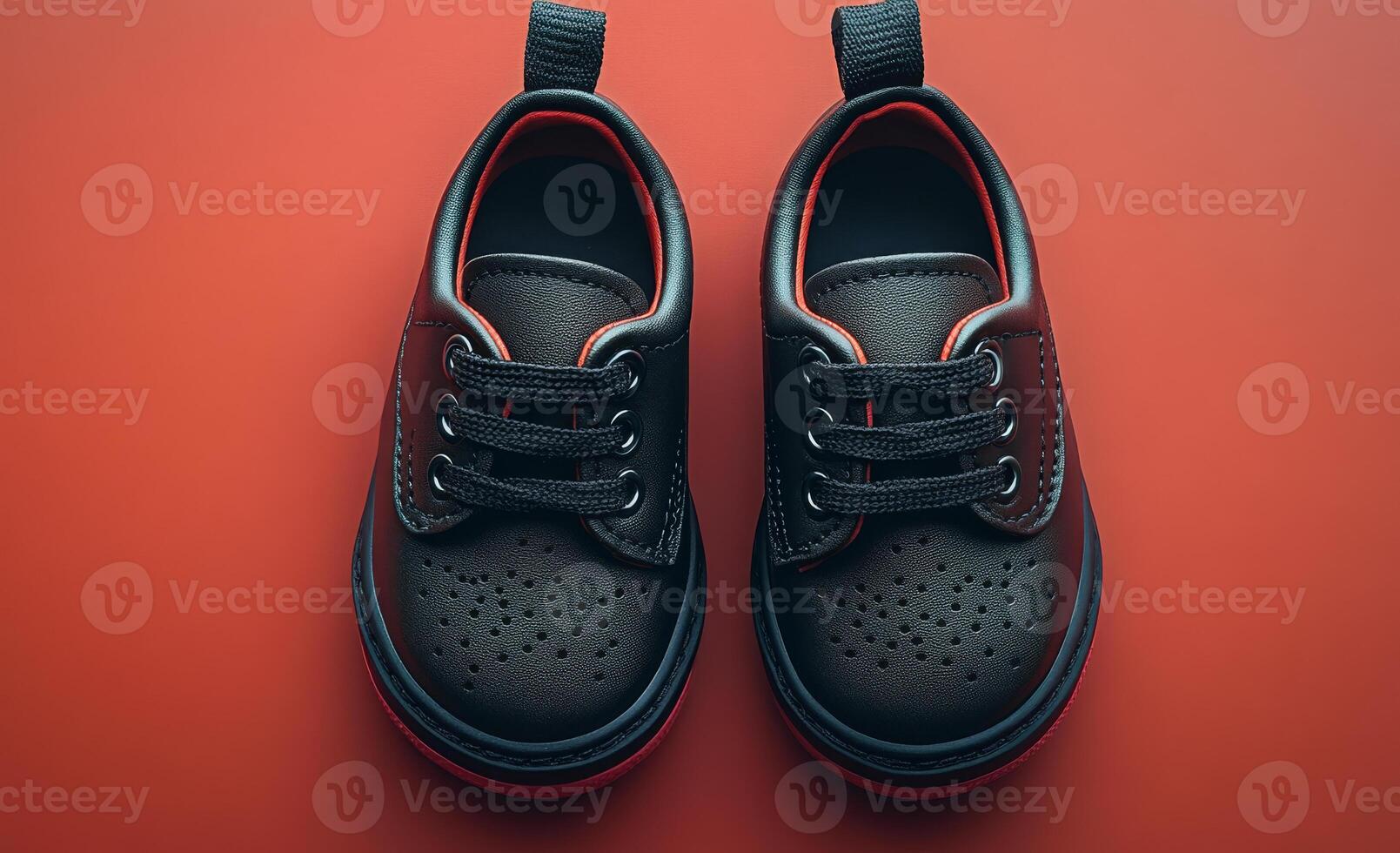 Two small black shoes with red laces are placed on a red surface. The shoes are positioned in such a way that they are almost fully visible, with their laces peeking out from under them photo