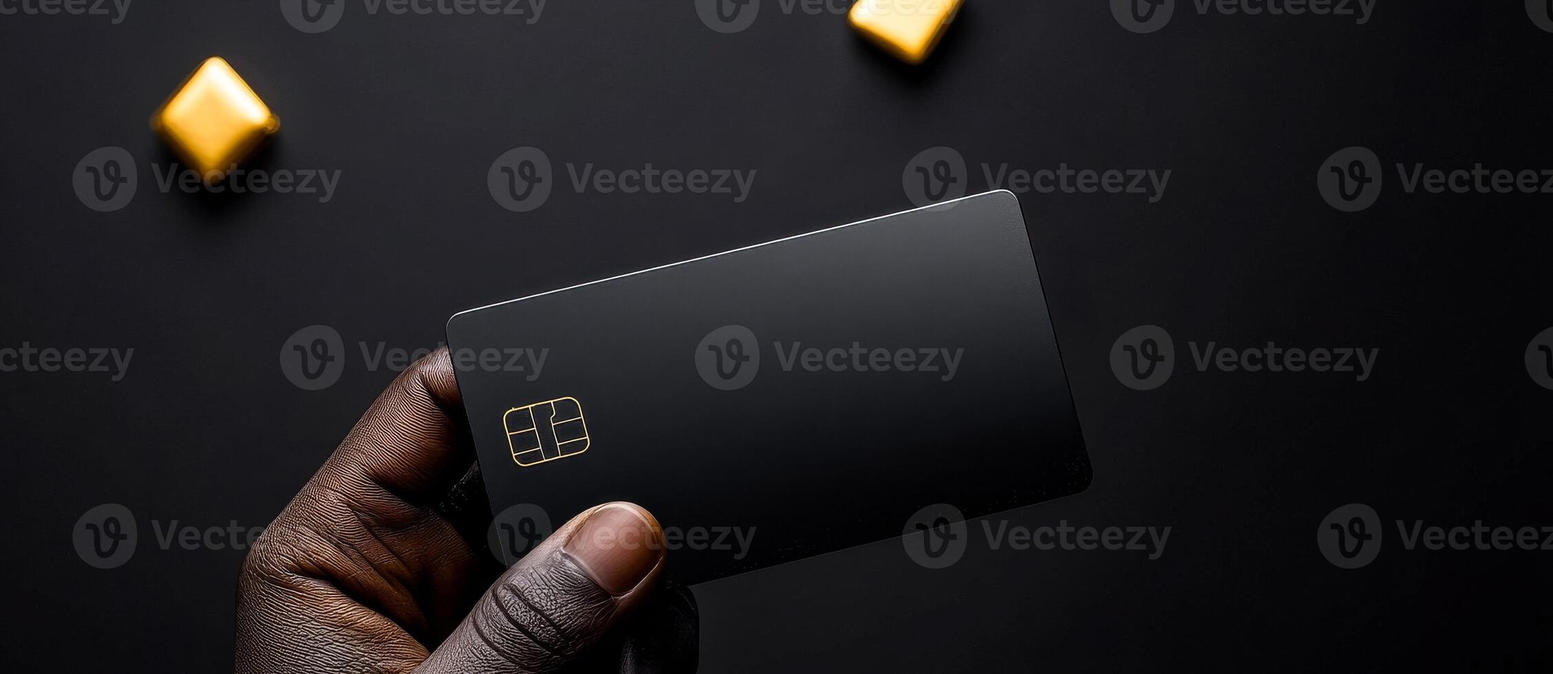 A person is holding a black credit card. The card is rectangular and has a gold border. The image has a dark background and a few yellow candies in the background photo