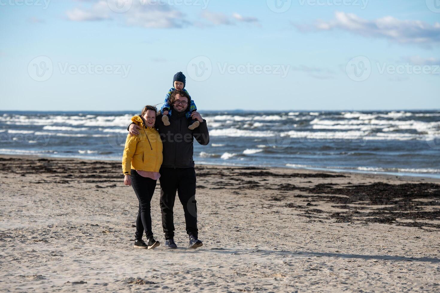 A happy family strolls along a chilly autumn beach, with their young son joyfully running ahead, creating lasting memories as the crisp wind and soothing waves embrace them in a moment of pure photo