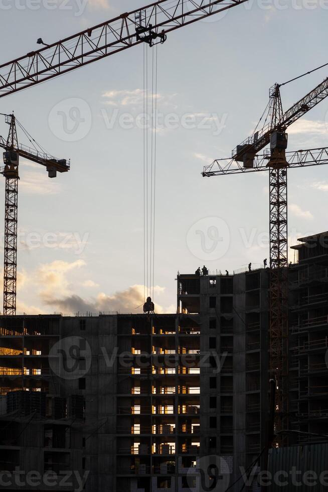Cranes operate at a construction site during sunset, with workers visible on the building's framework as daylight fades photo