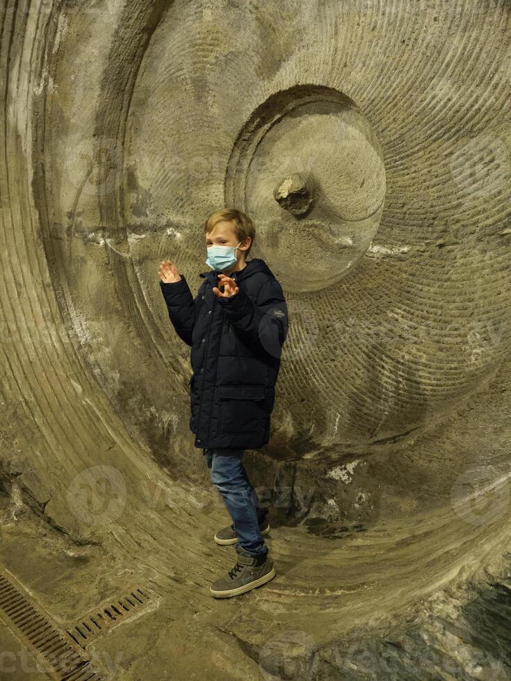 A child wearing a face mask stands in front of a large concrete wall photo