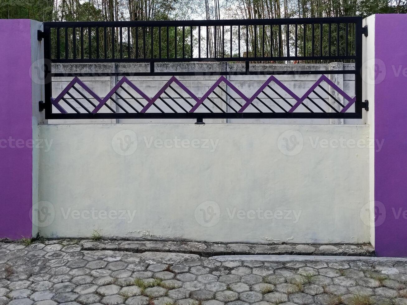 A modern fence with a purple accent wall and pillars. The fence features a geometric design and is situated in front of a bamboo grove. photo
