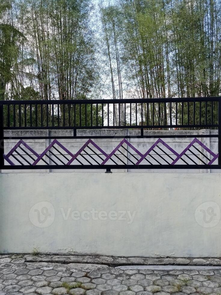 A modern fence with a purple accent wall and pillars. The fence features a geometric design and is situated in front of a bamboo grove. photo