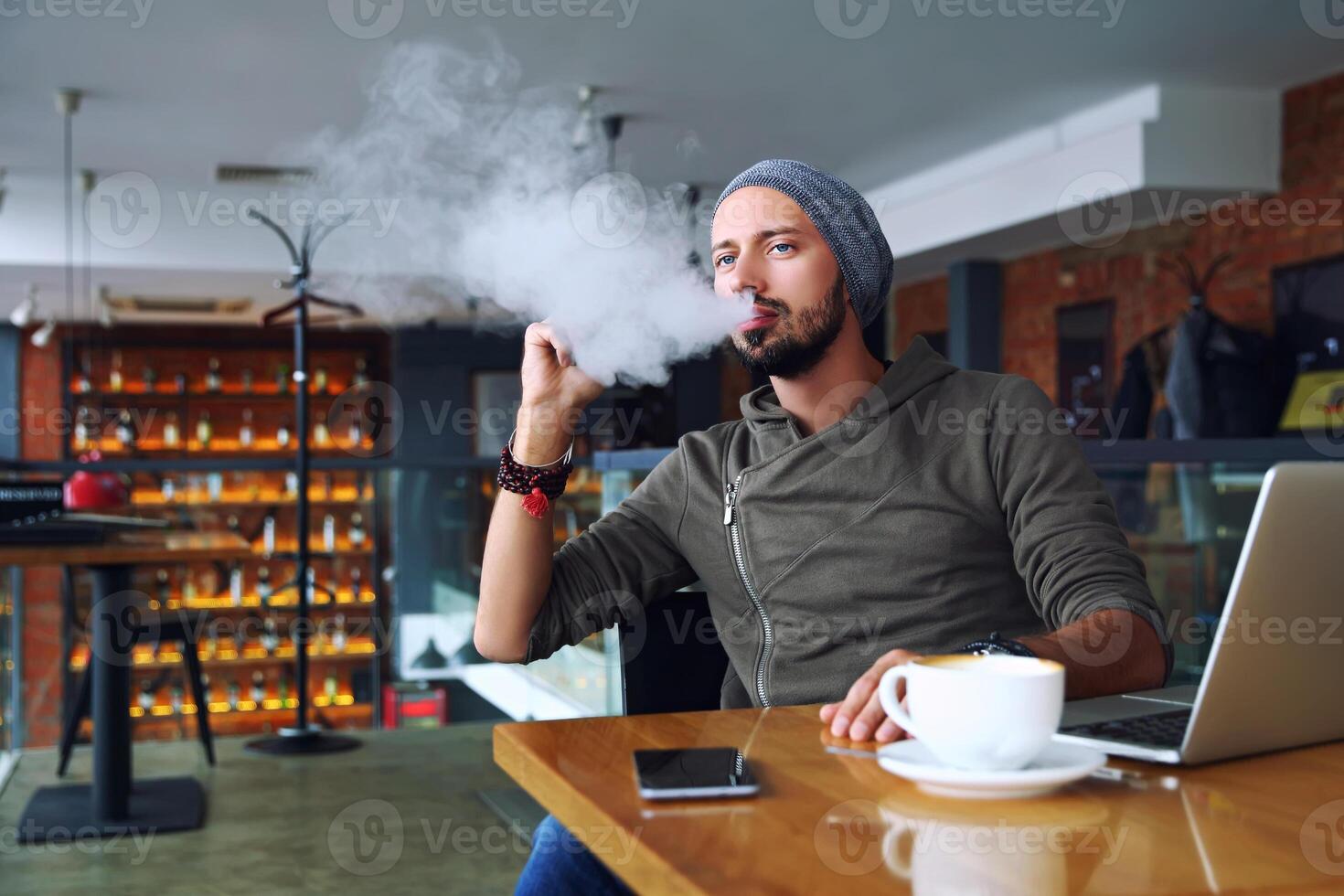 Young handsome hipster man with beard sitting in cafe with a cup of coffee, vaping and releases a cloud of vapor. Working at laptop and having a little break. With copy space. photo