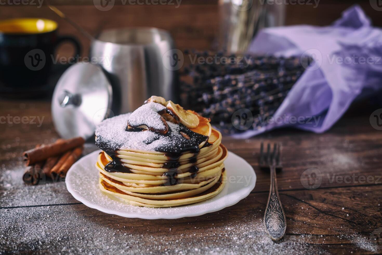 Stack of delicious pancakes with chocolate, honey, slices of banana and castor sugar on plate on wooden background, cinnamon sticks and bunch of lavender. photo